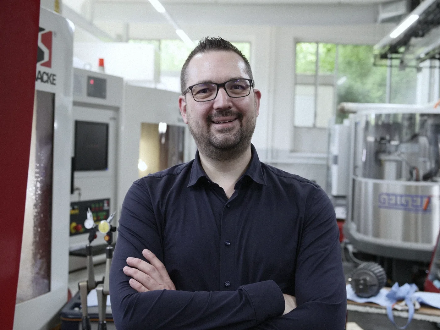 Robin Steinheimer steht mit verschränkten Armen lächelnd in einer Fertigungshalle vor CNC Maschinen
